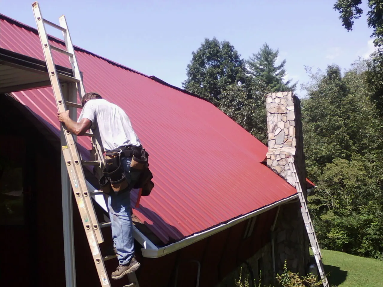Roofer on a ladder beside a red metal roof and stone chimney — Soffit & Fascia Repair by Roofing Brothers in Coolville